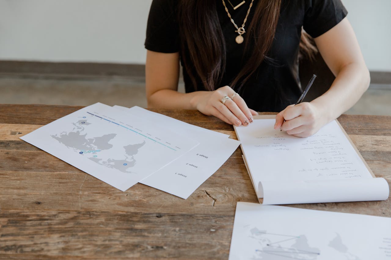 A woman in a black shirt takes notes at a wooden table with maps and papers.