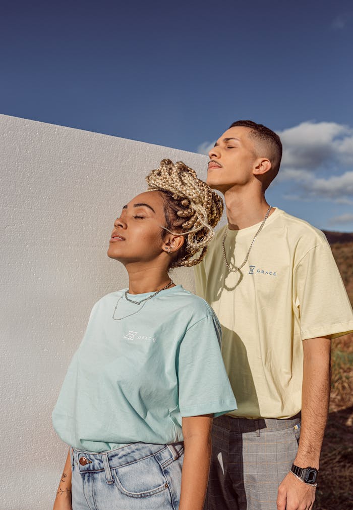 A stylish couple with closed eyes standing outdoors against a white wall in pastel t-shirts.